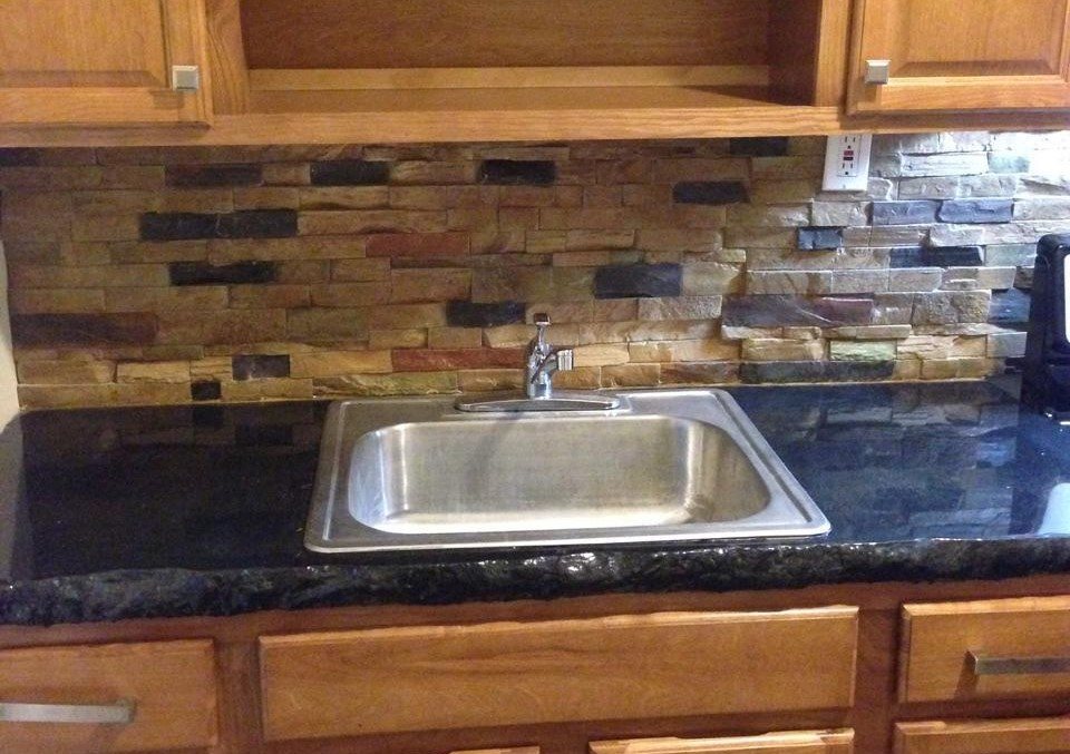 A kitchen backsplash of warm gold and rust stacked stone with scattered dark stones, above a black granite countertop with a stainless sink and oak cabinets.