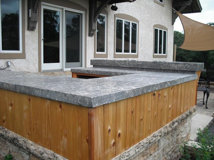 An outdoor kitchen bar with cedar cladding and a thick grey and white speckled concrete countertop, outside a stucco house.