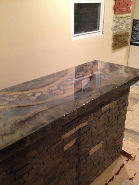 A bar with a glossy grey and cream concrete top containing a river of embedded stones, on a base faced with dark stacked stone panels.