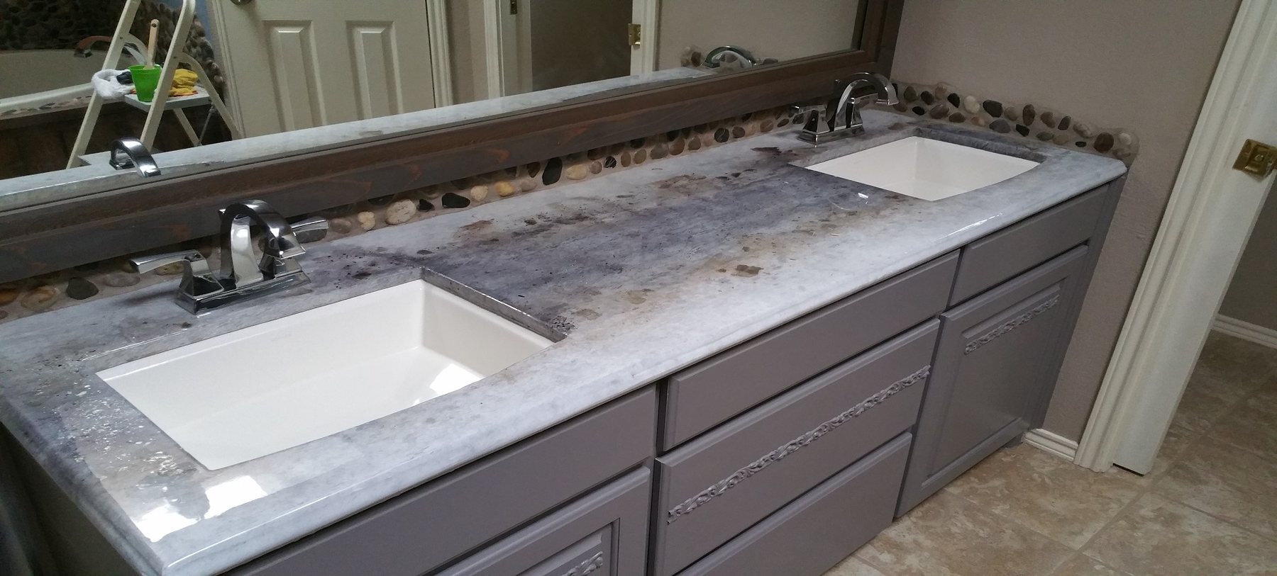 A long bathroom double vanity with a grey and white marbled concrete top, two white undermount basins, chrome taps and a backsplash of rounded river stones.