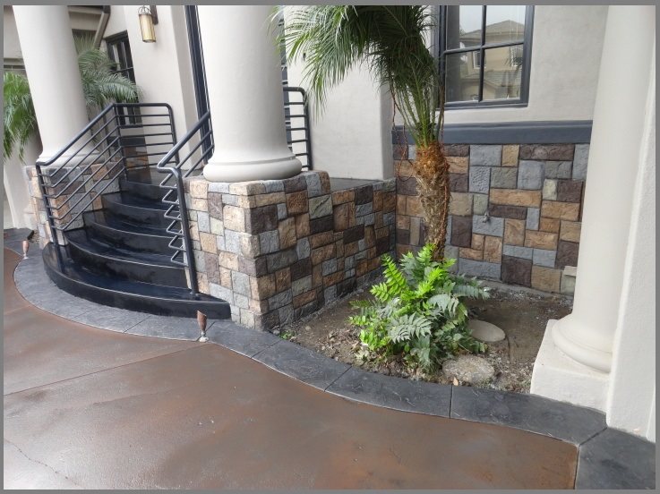 The entrance of a house with white columns and stone veneer, with a concrete walkway edged by a dark grey stamped border curving around black steps.