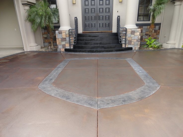 A wide concrete entry court coloured warm brown with a grey stamped border framing a fan shape in front of black steps and a dark front door.
