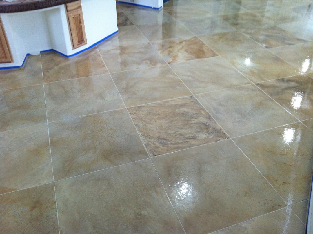A glossy interior concrete floor scored into large squares and stained in cream, tan and grey marbled tones, next to a kitchen island.