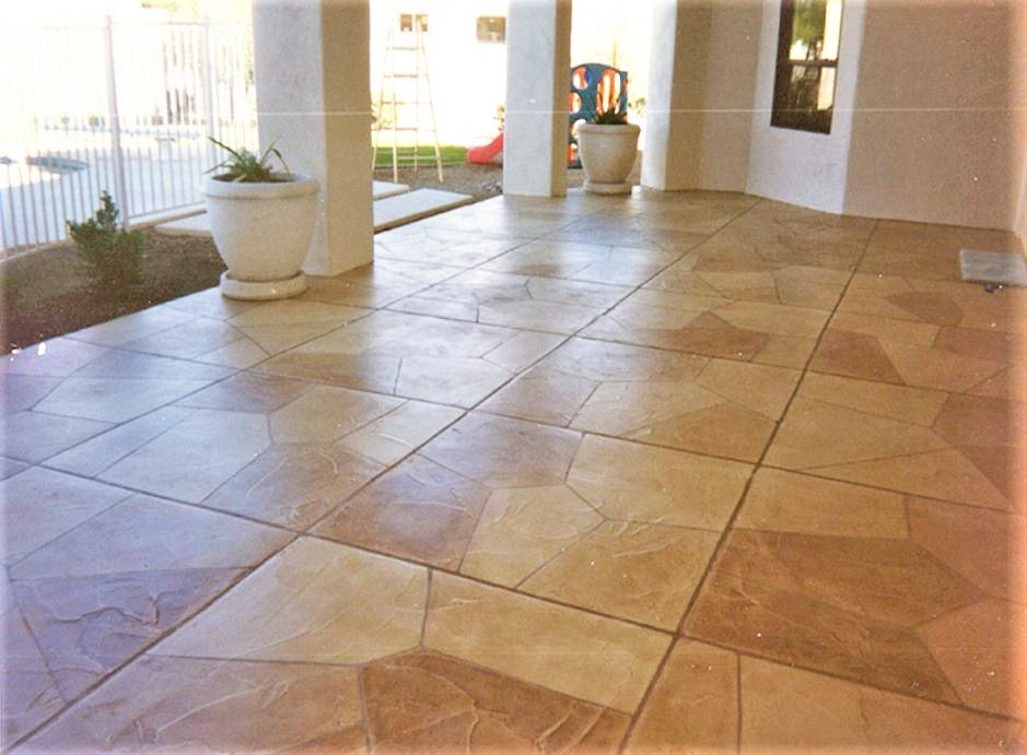 A covered patio finished in gold and tan concrete stamped as large random flagstone, with white columns and planters, beside a pool fence.
