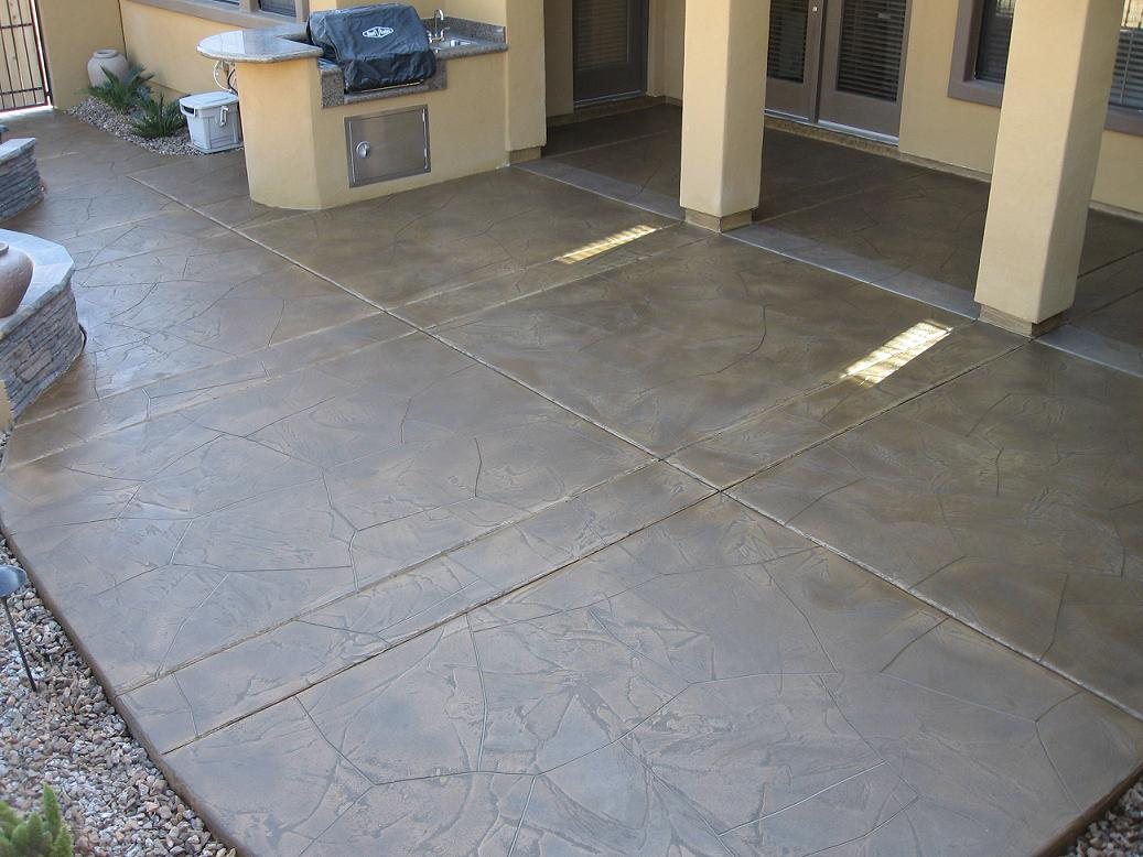 A large brown concrete patio stamped as random flagstone, running past a built-in outdoor kitchen with a grill and stone-clad columns.