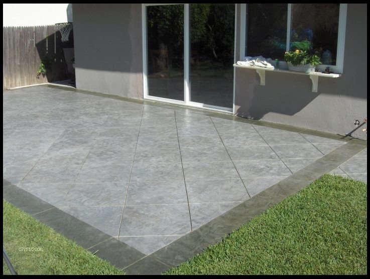 A back patio finished in pale grey concrete scored into large diagonal tiles with a darker grey border, beside a house with sliding glass doors.