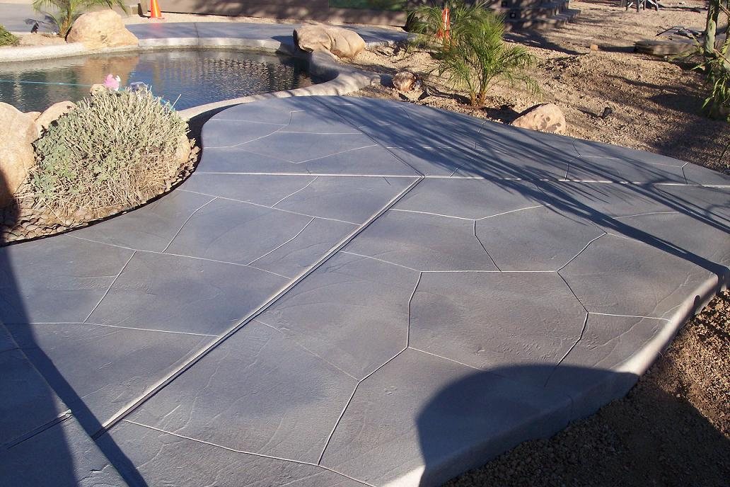 A grey concrete pool deck stamped in a random flagstone pattern, curving around a swimming pool with desert planting and boulders.