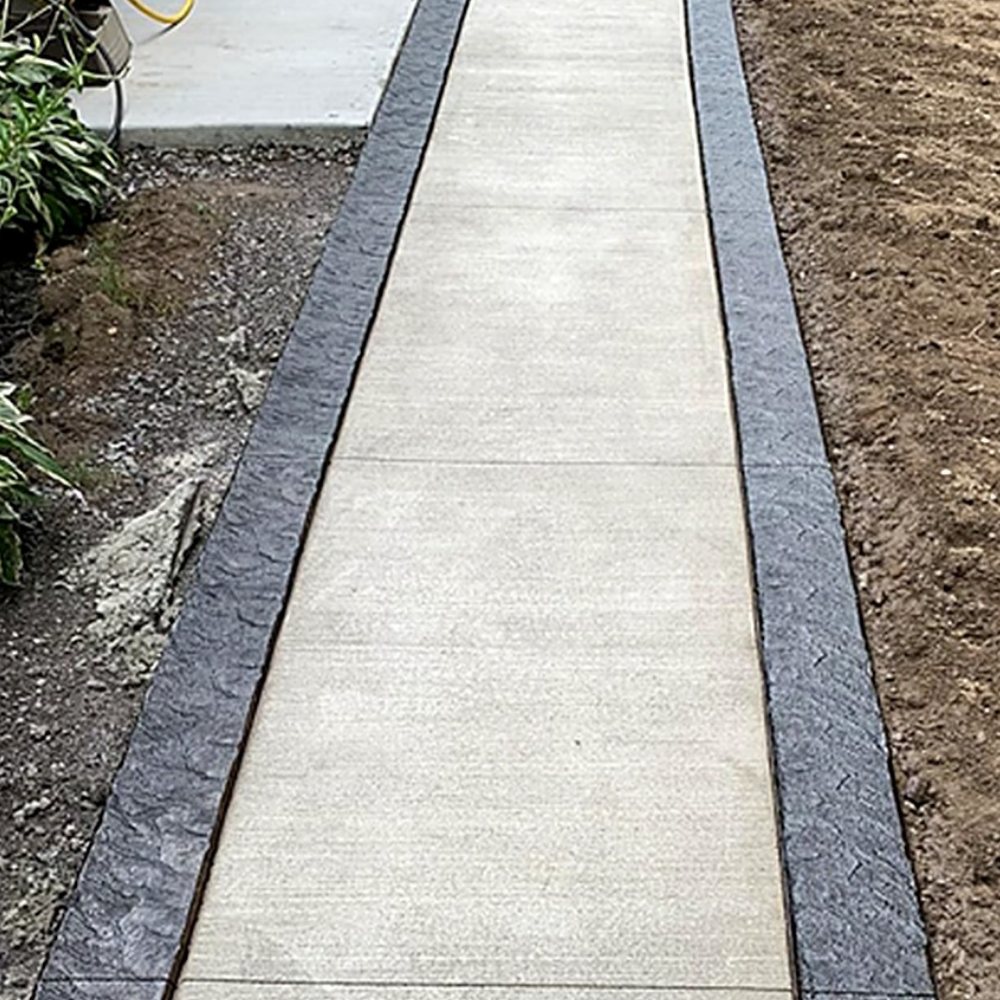 A newly poured concrete walkway with a smooth pale centre and dark Weathered Flagstone textured bands running along both edges, with bare soil either side.