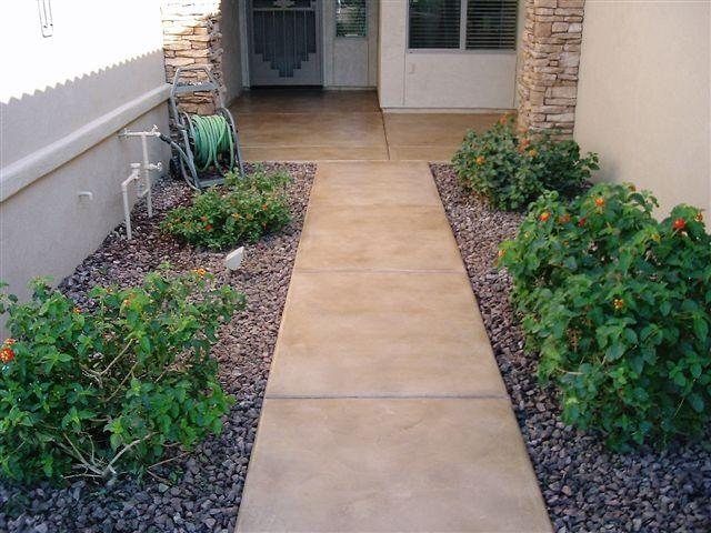 A narrow side walkway stained warm tan and scored into long panels, running between desert gravel planting beds beside a stucco house.