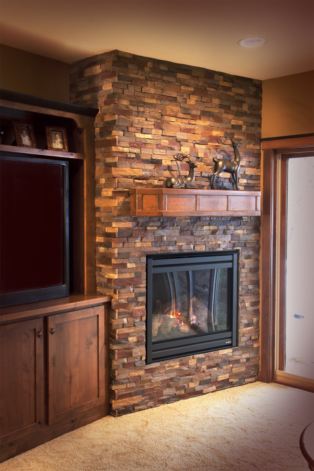 A floor-to-ceiling fireplace and media wall clad in warm brown, gold and slate stacked stone panels, with a timber mantel and a lit gas fire.