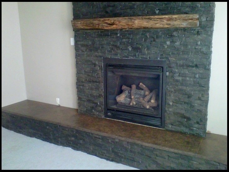 A fireplace clad in dark green and grey stacked stone panels with a rustic timber mantel beam and a long brown concrete hearth.