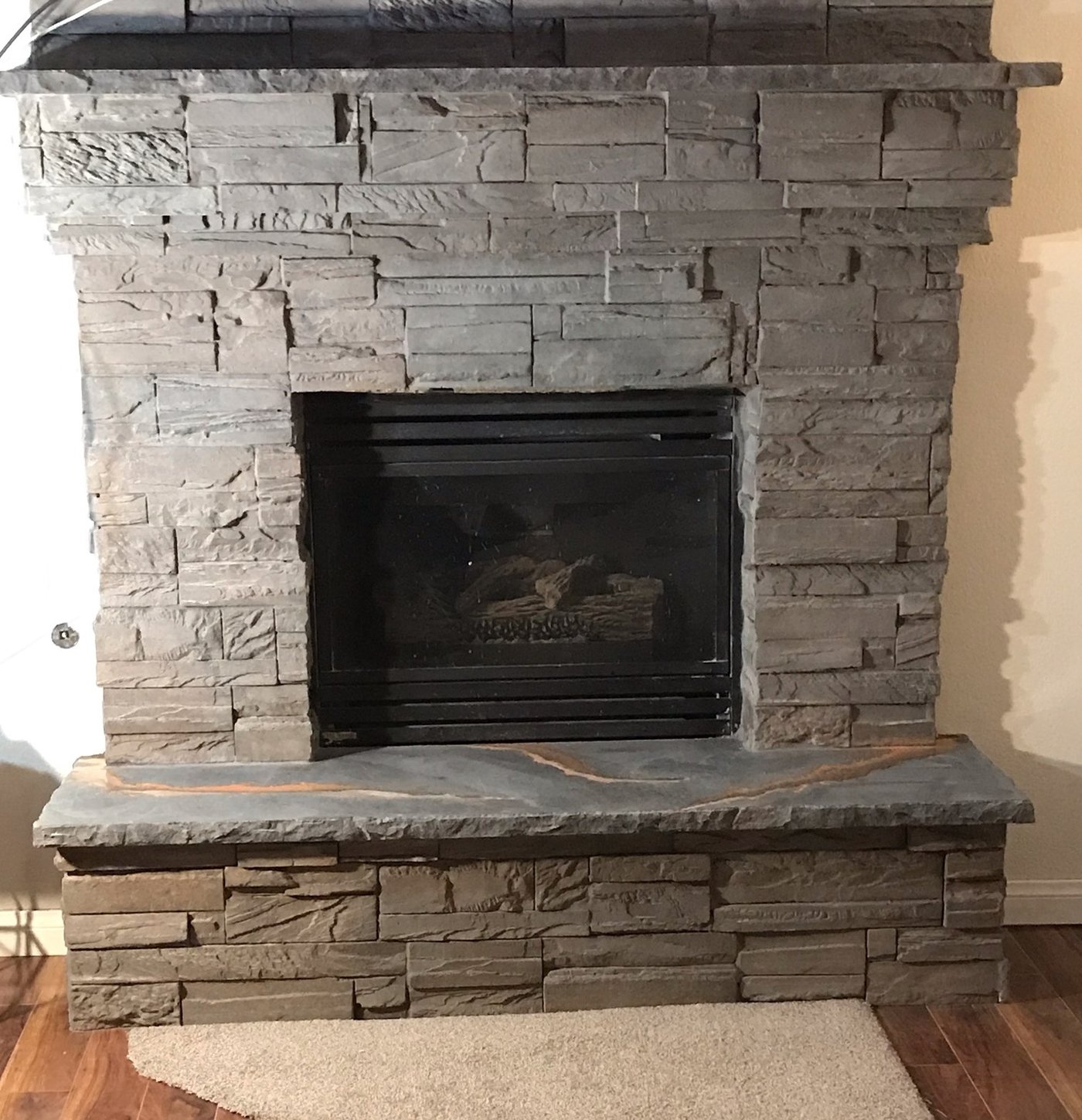 A floor-to-ceiling fireplace clad in grey stacked stone panels with a cast concrete mantel shelf and a grey hearth slab veined with rust, on a wood floor.
