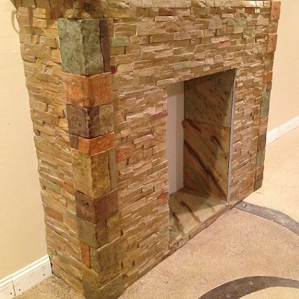 A freestanding fireplace surround clad in multi-coloured green, gold and rust stacked stone panels, showing the mitred corner return.