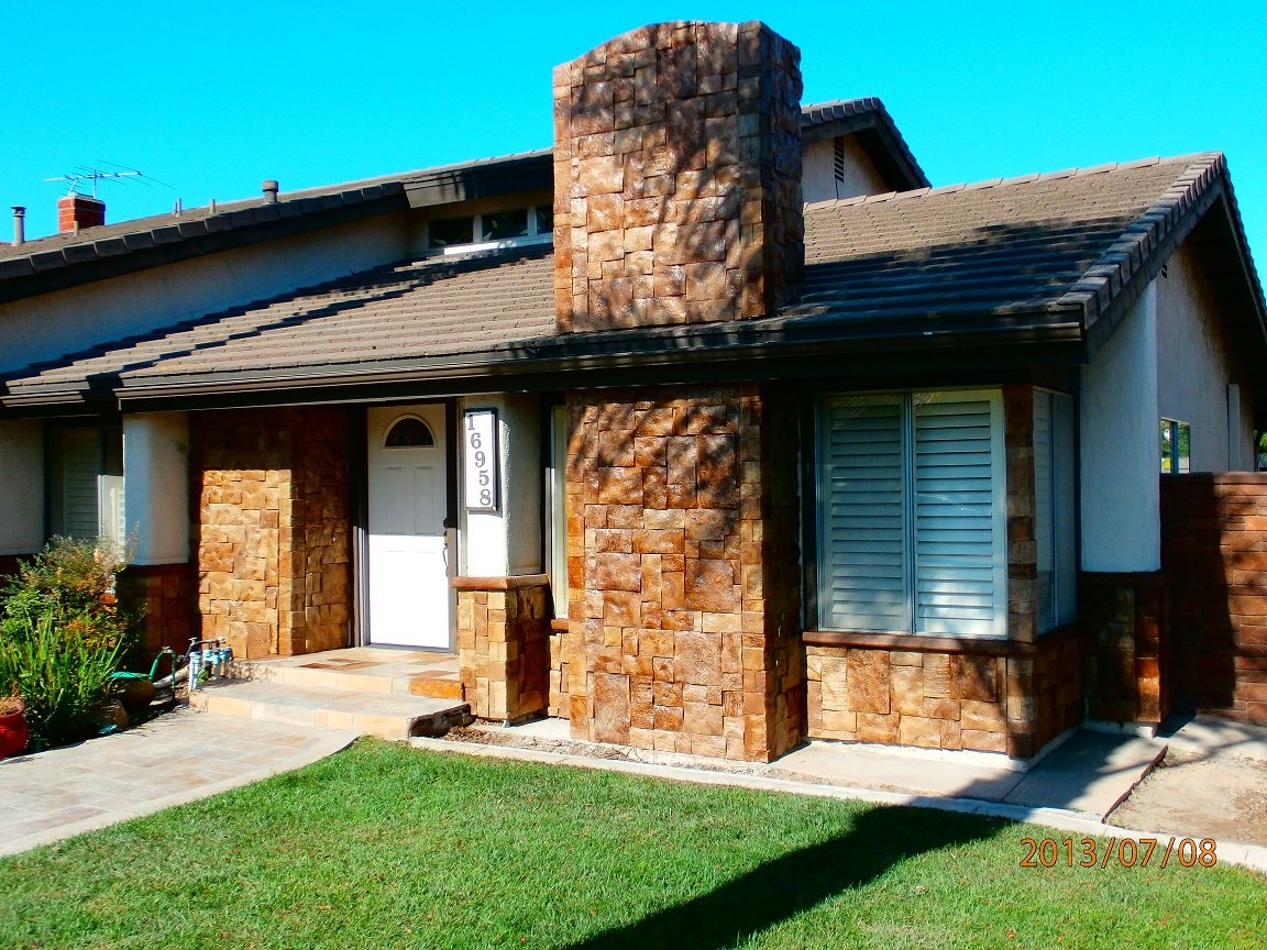 The front of a single-storey house with a tall chimney breast and two entry columns clad in gold and amber stacked stone panels, above a green lawn.