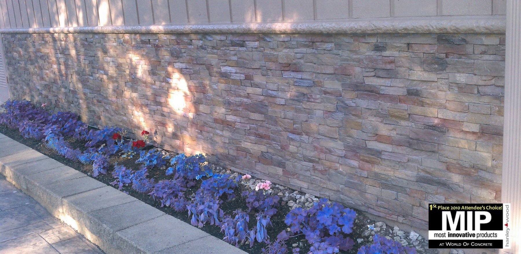 A long low garden boundary wall clad in pale multi-coloured stacked stone panels with a cast concrete cap, above a bed of blue and purple flowers.