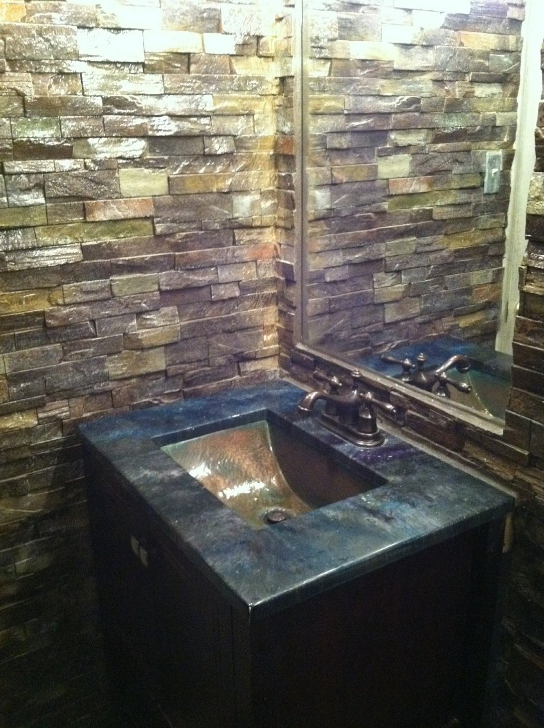 A powder room with walls clad in warm multi-coloured stacked stone, a dark vanity with a blue-green marbled concrete top and a hammered copper undermount basin.