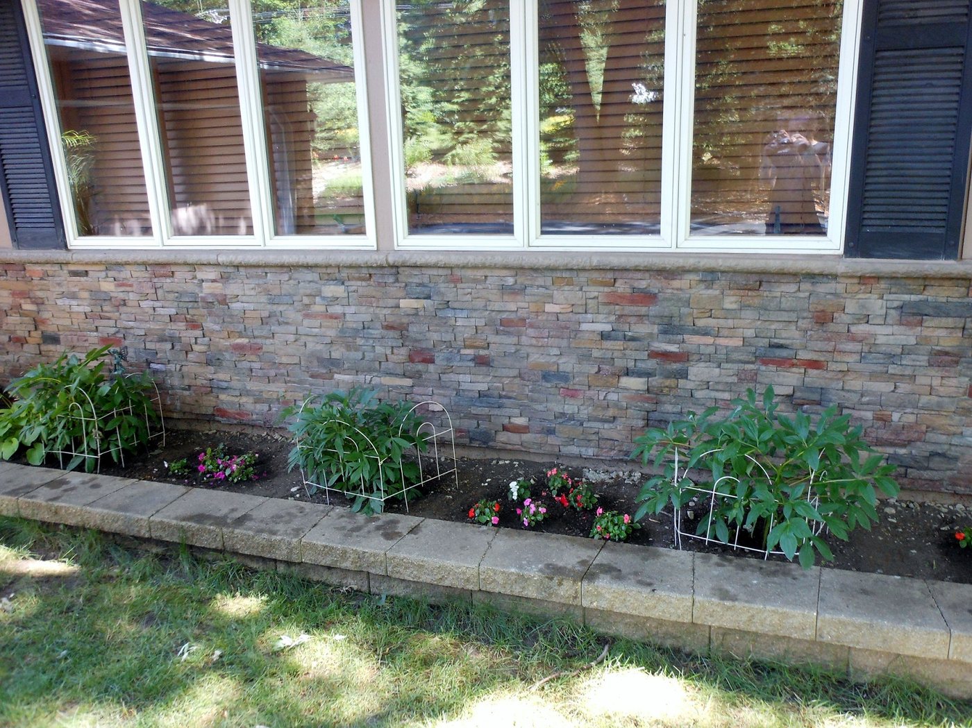 A long house wainscot below a run of windows, clad in stacked stone panels mixing grey, rust and ochre, above a raised planting bed with flowers.